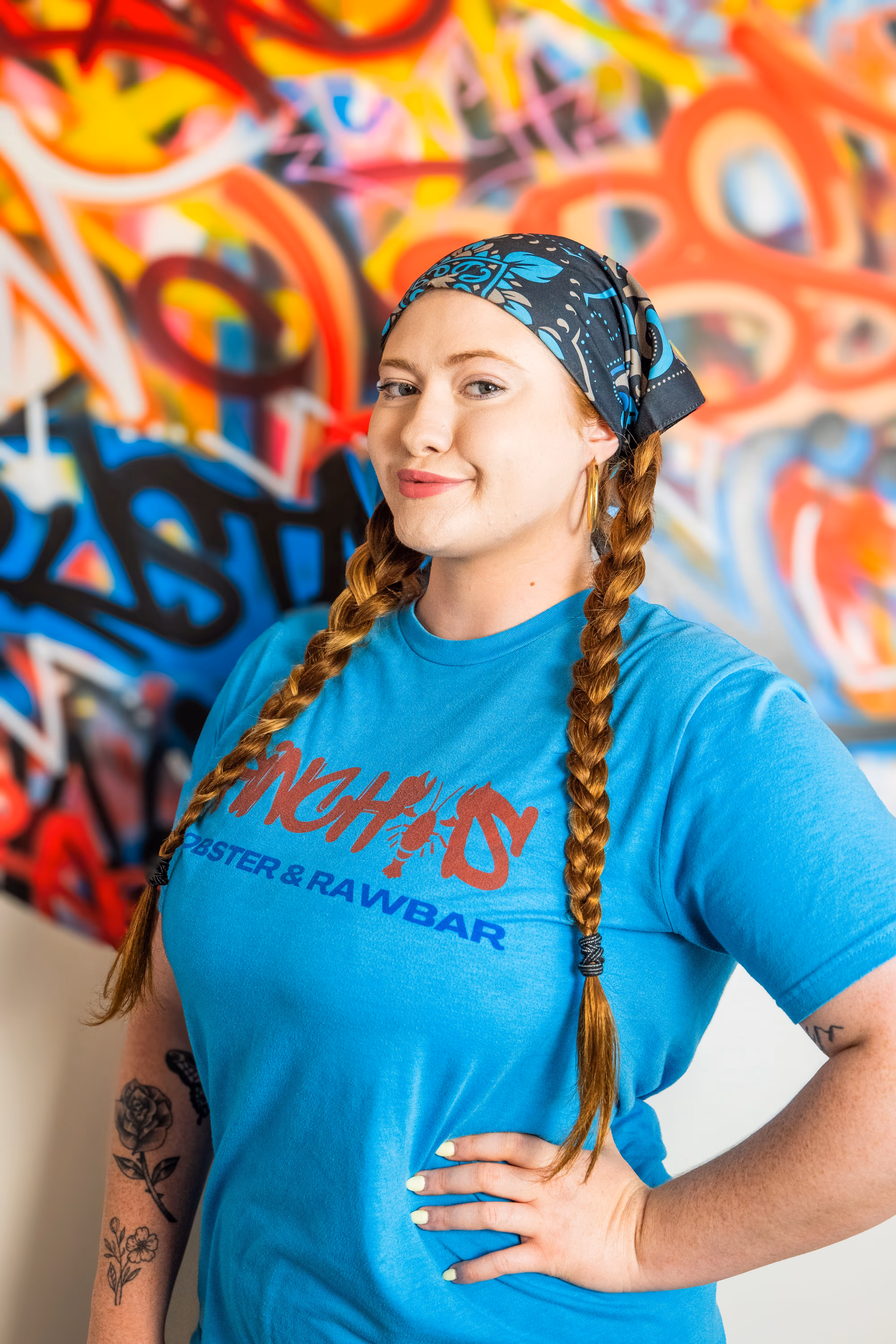 Woman with braided red hair and blue bandana, wearing a blue T-shirt with tattoos on her arm, in front of graffiti.