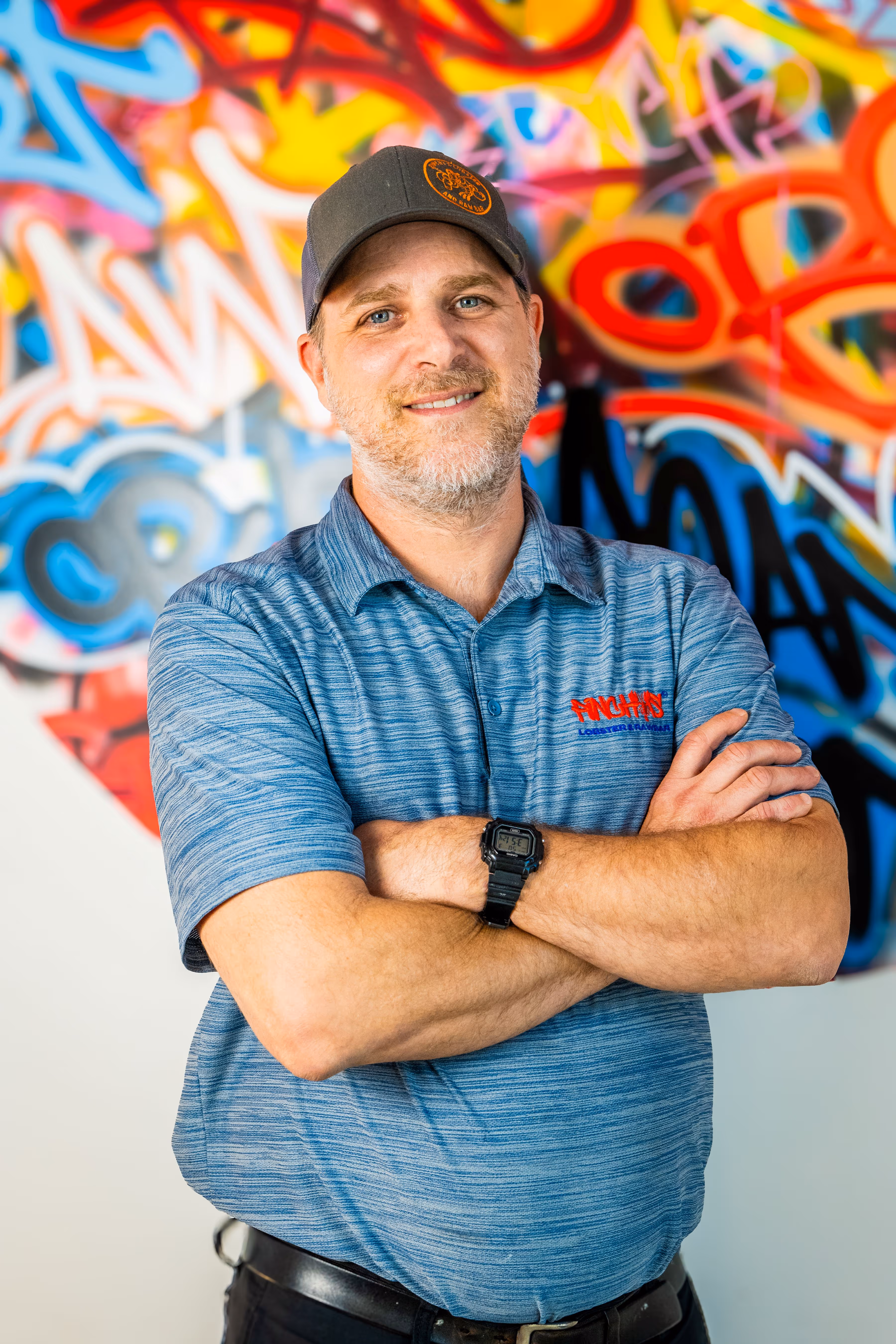 Man with a beard in a blue shirt and cap standing with arms crossed in front of colorful graffiti wall.