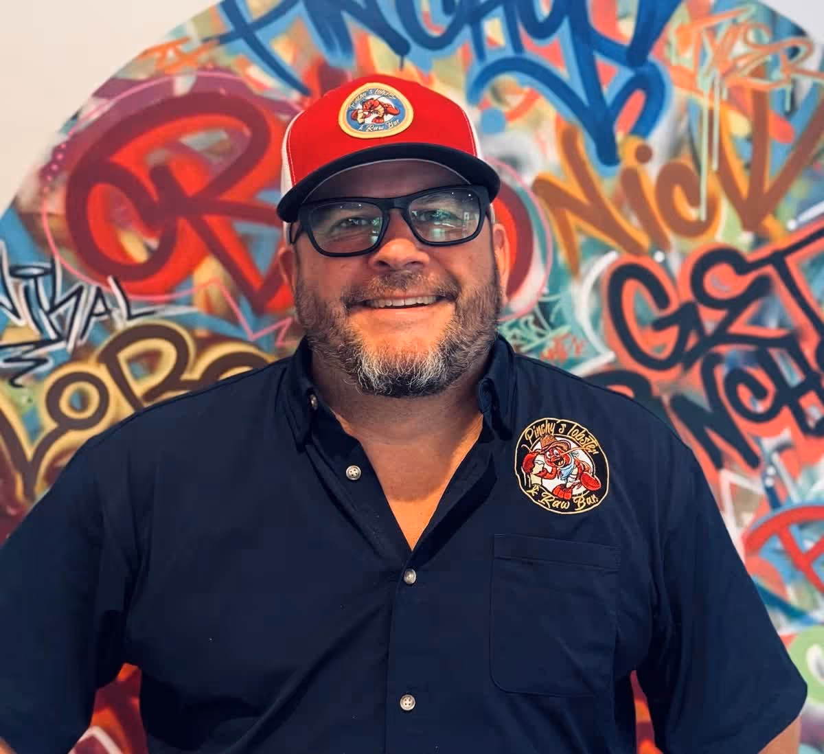 Smiling man with glasses wearing a red cap and black shirt with Pinchy's Lobster Raw Bar logo.