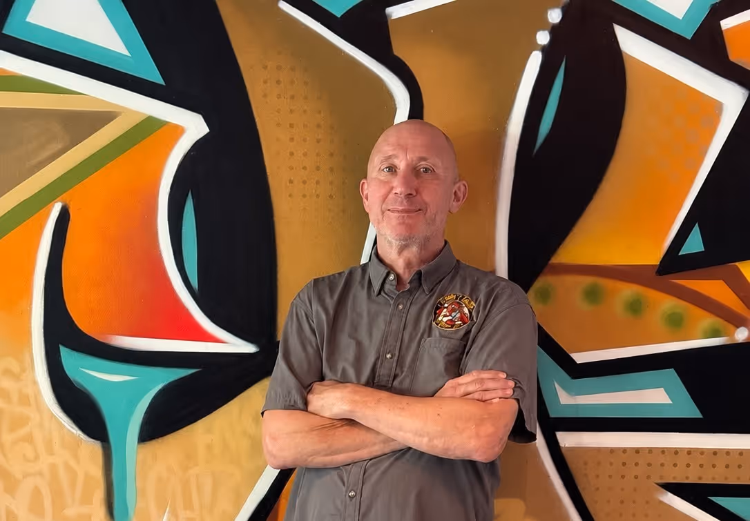 Smiling bald man with crossed arms in gray shirt stands before colorful abstract graffiti wall.