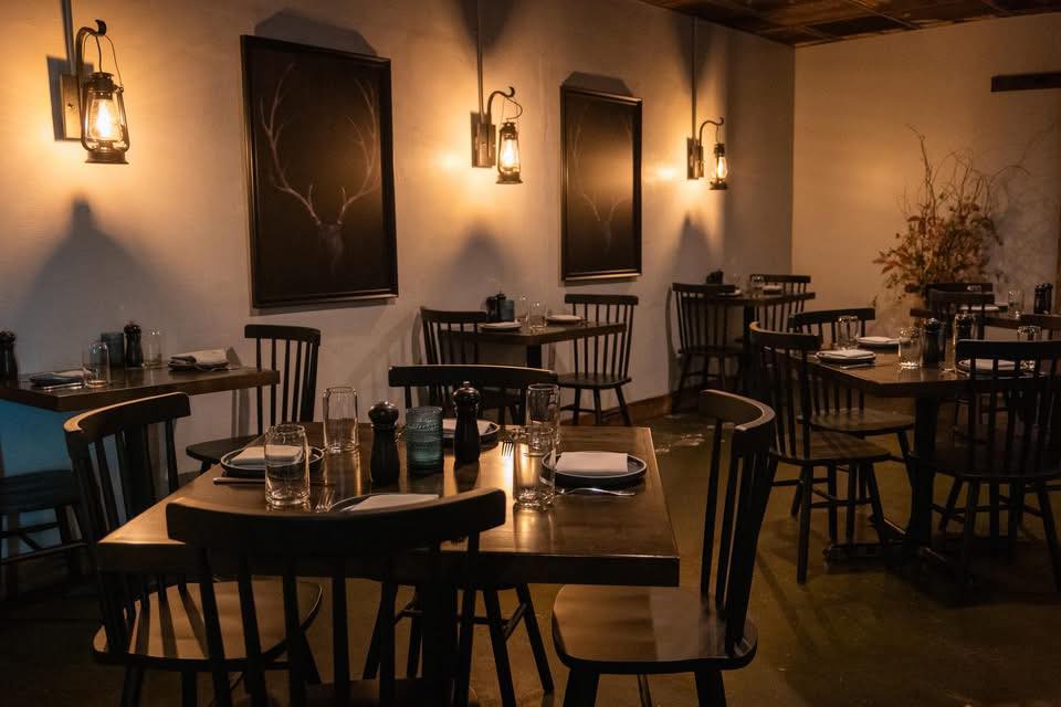 Cozy dimly lit restaurant with wooden tables, chairs, lantern wall lights, and deer antler paintings.