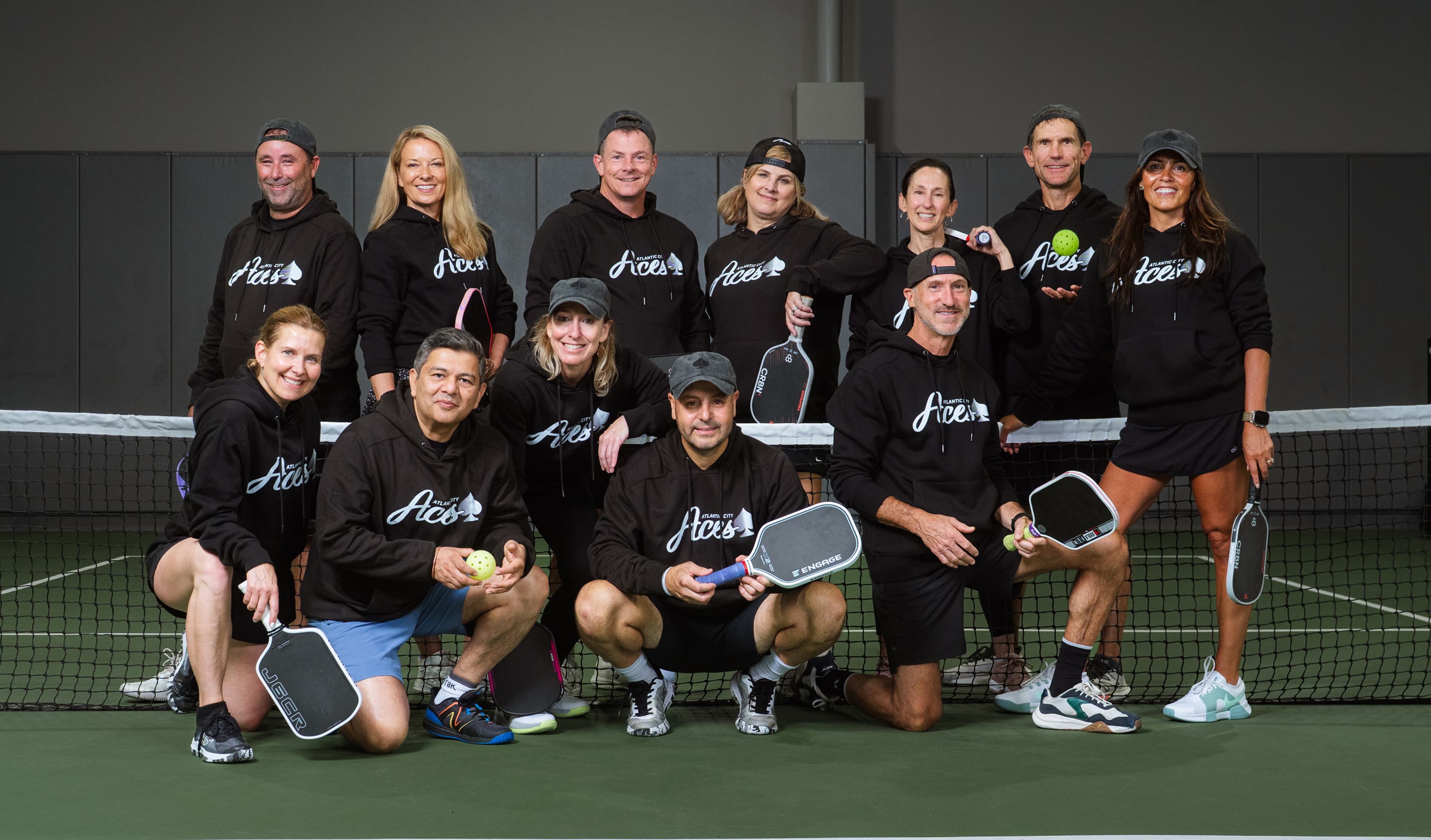 Thirteen people in black Atlantic City Aces hoodies pose on a pickleball court holding paddles and balls.