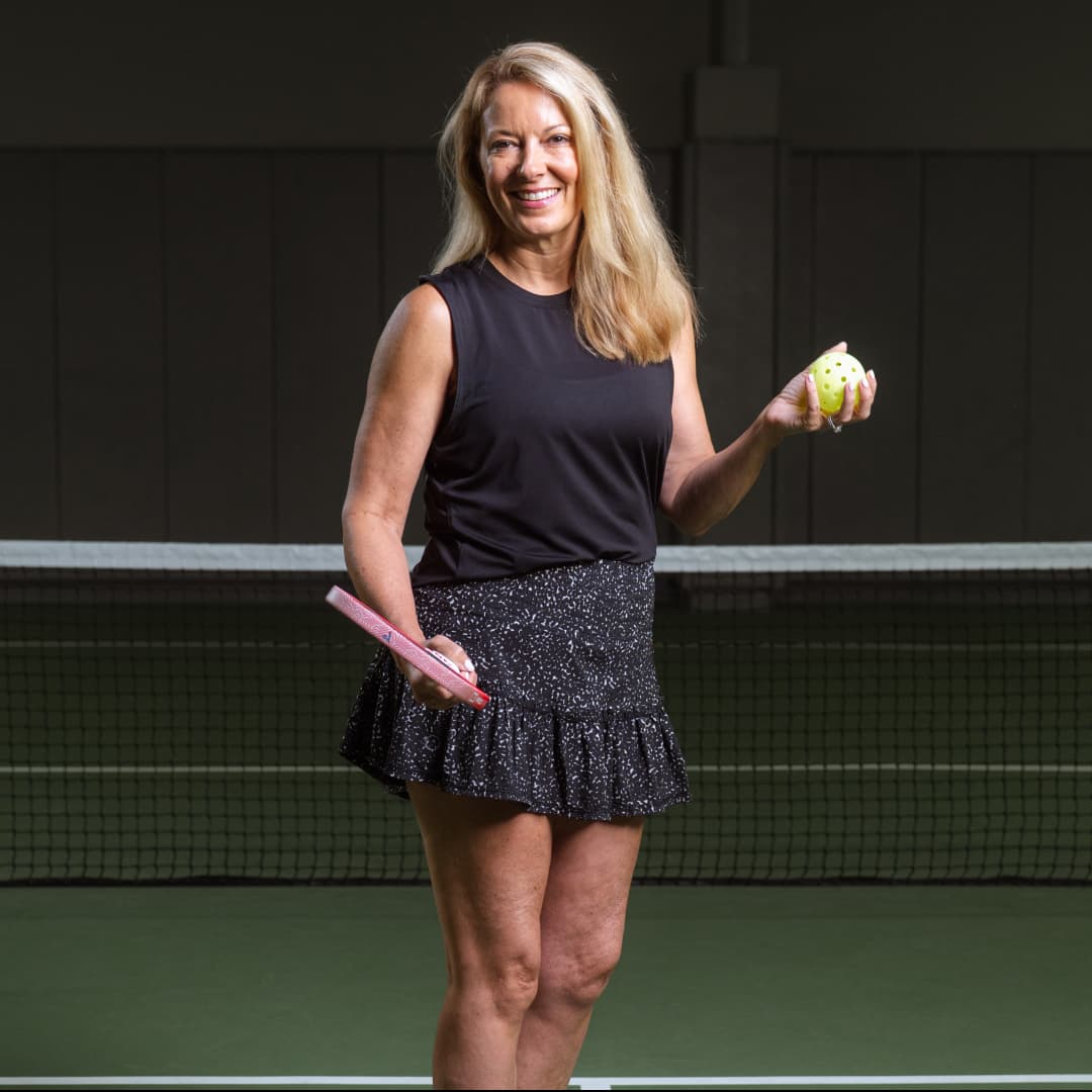 Fun profile photo of Jen Gelwas wearing a black tank top and skirt holding a paddle in one hand and pickleball in the other