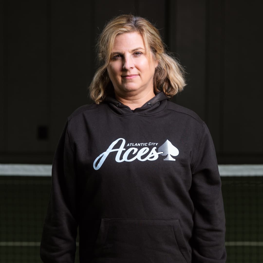Photo of Sheri Courter waste up wearing an Atlantic City Aces black hoodie standing in front of a pickleball net. 