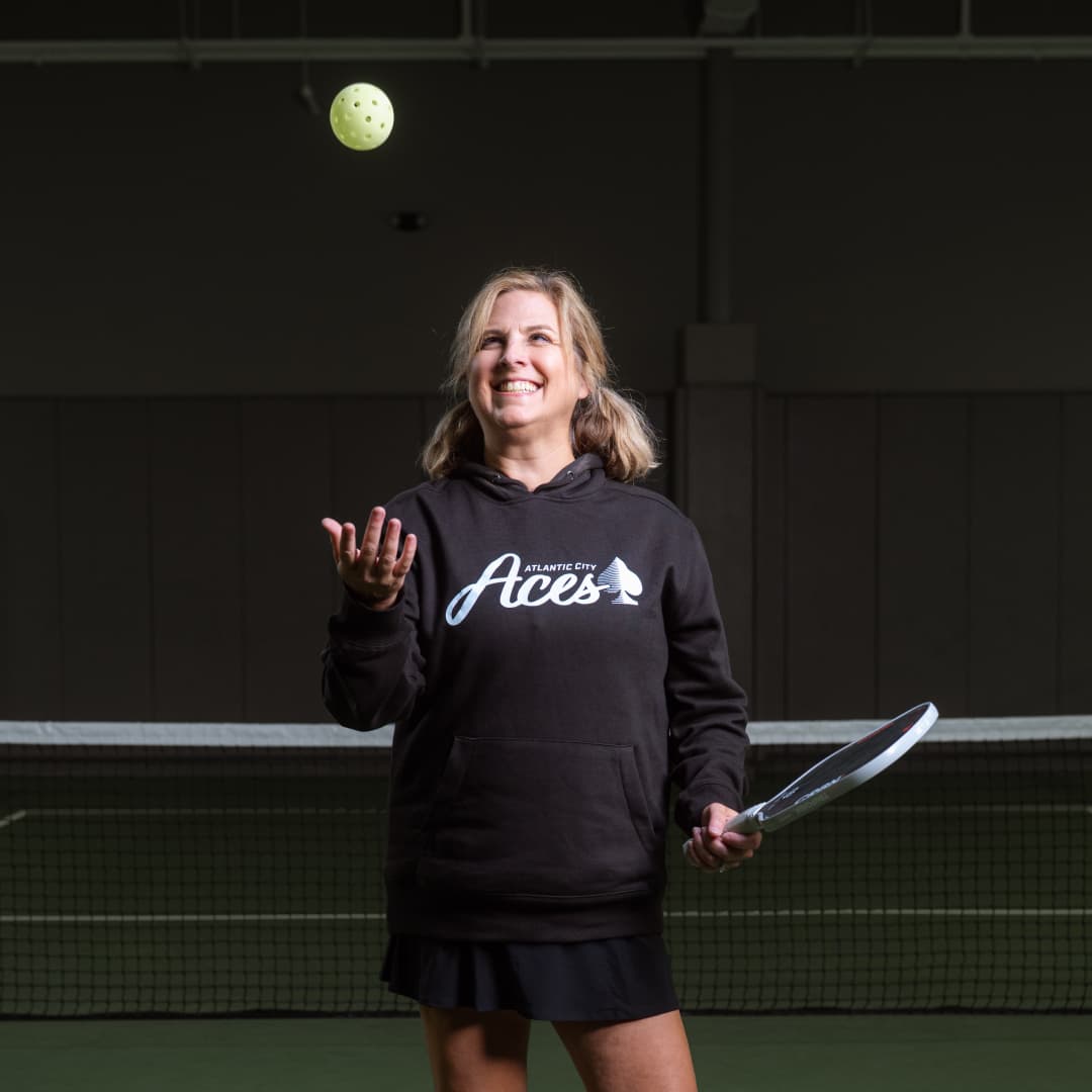 Photo of Sheri Courter wearing a Atlantic City Aces black hoodie tossing up a pickball with her right arm. 