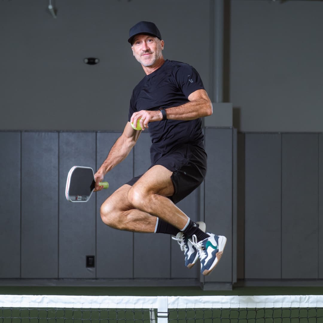 Picture of Hugh Laroque jumping above a pickleball net wearing a black hat and shirt while holding a pickleball paddle in right hand.
