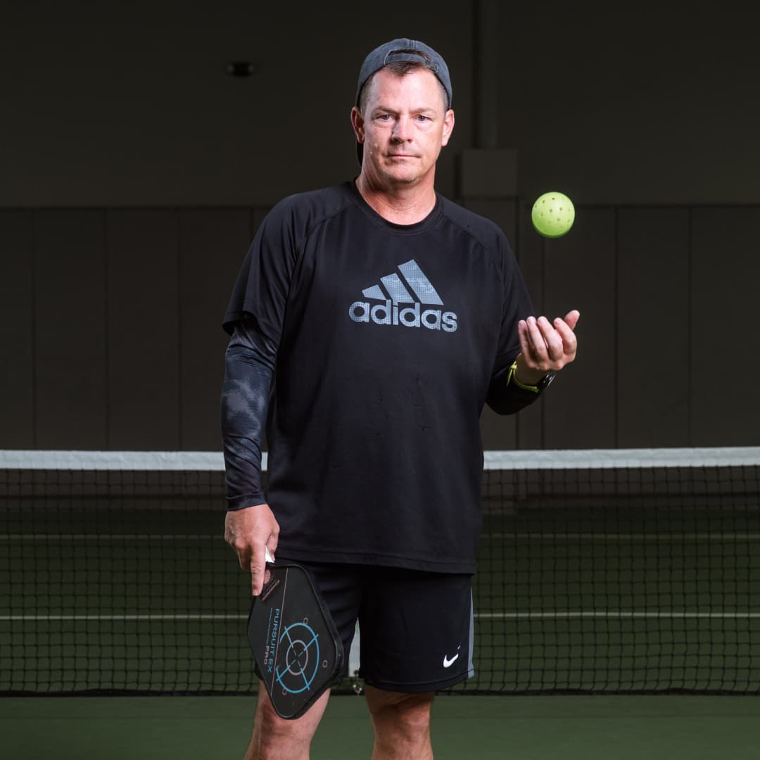 Profile picture of Chris Wilton wearing a black hat and black Adidas branded shirt catching a pickleball with left hand.