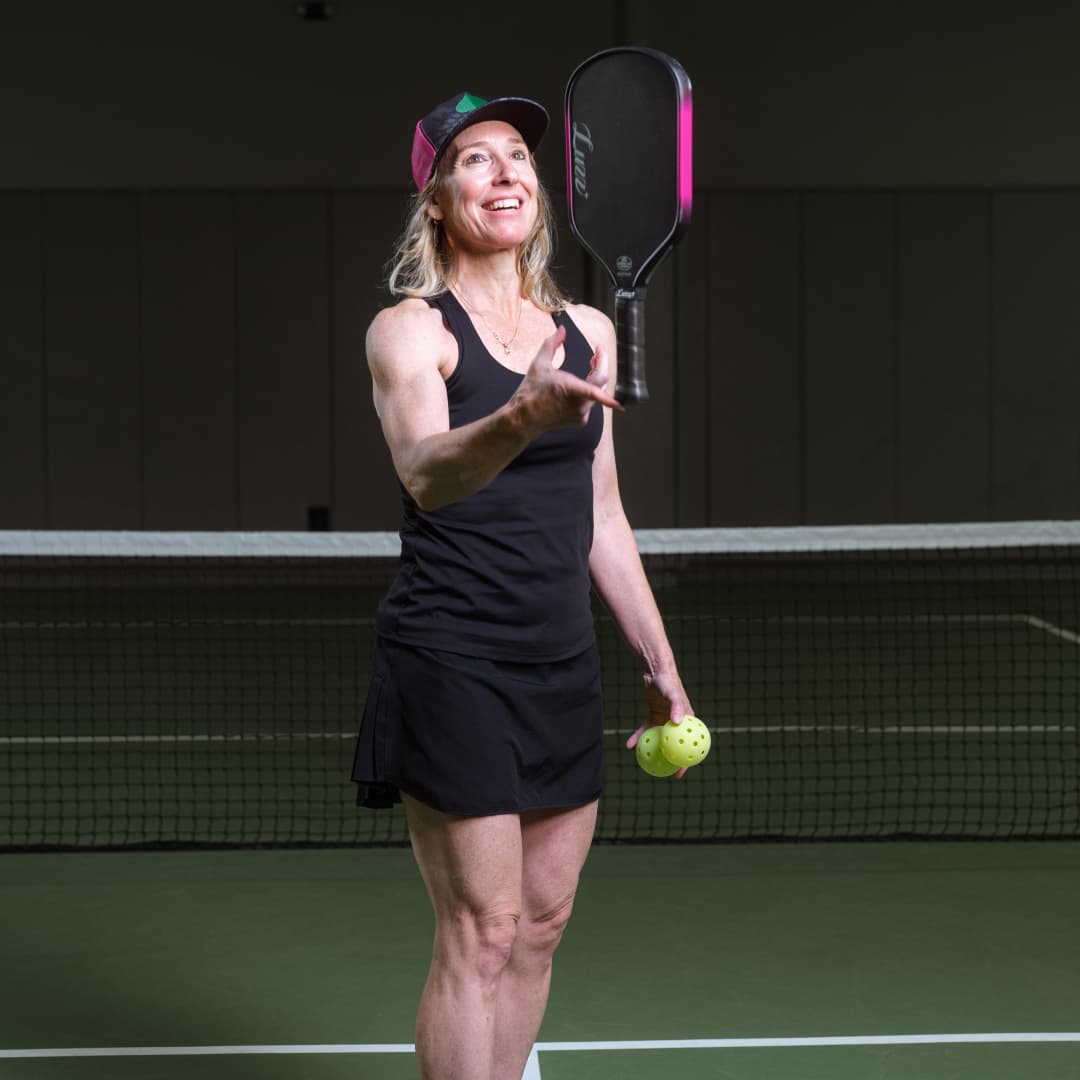 Profile picture of Jill Belyea wearing a black Atlantic City Aces hat and a black tanktop balancing a pickleball paddle with right hand.