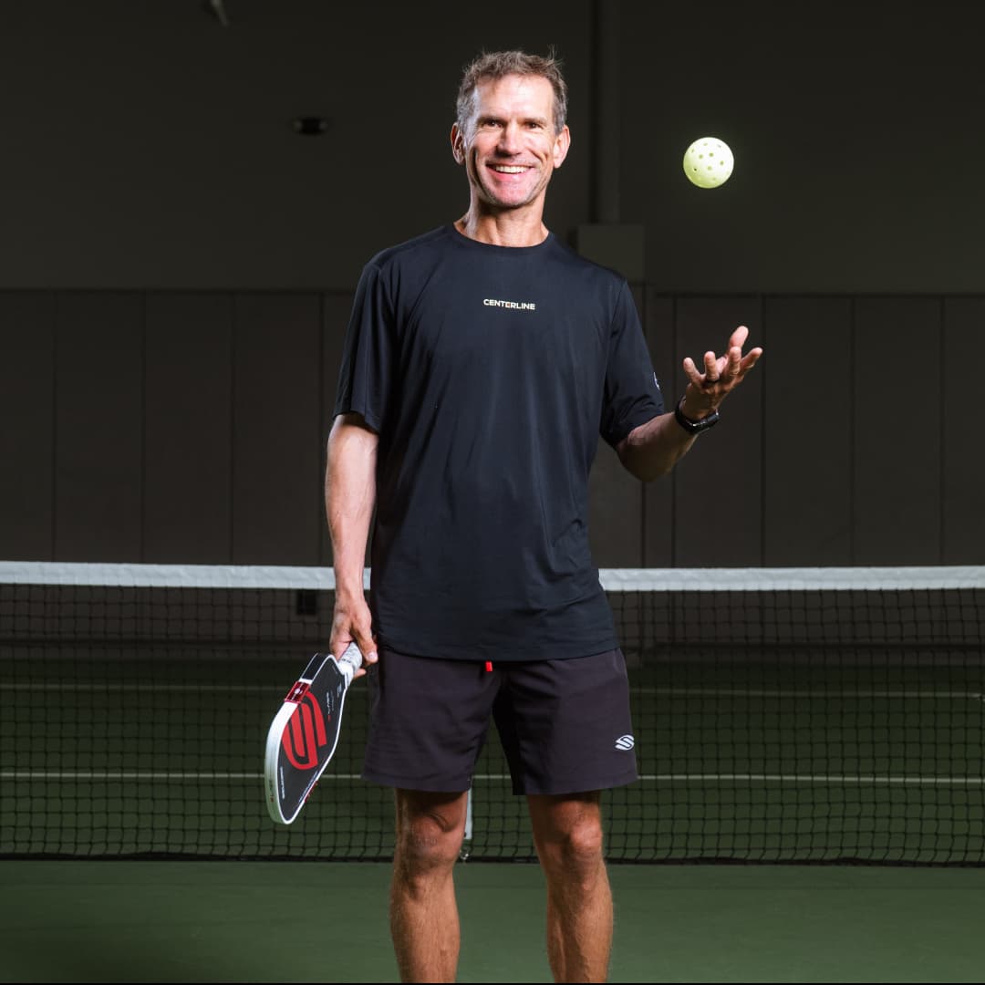 Profile picture of Paul Salmela wearing a black Centerline t-shirt and black shorts catching a pickleball with left hand and holding paddle with right.