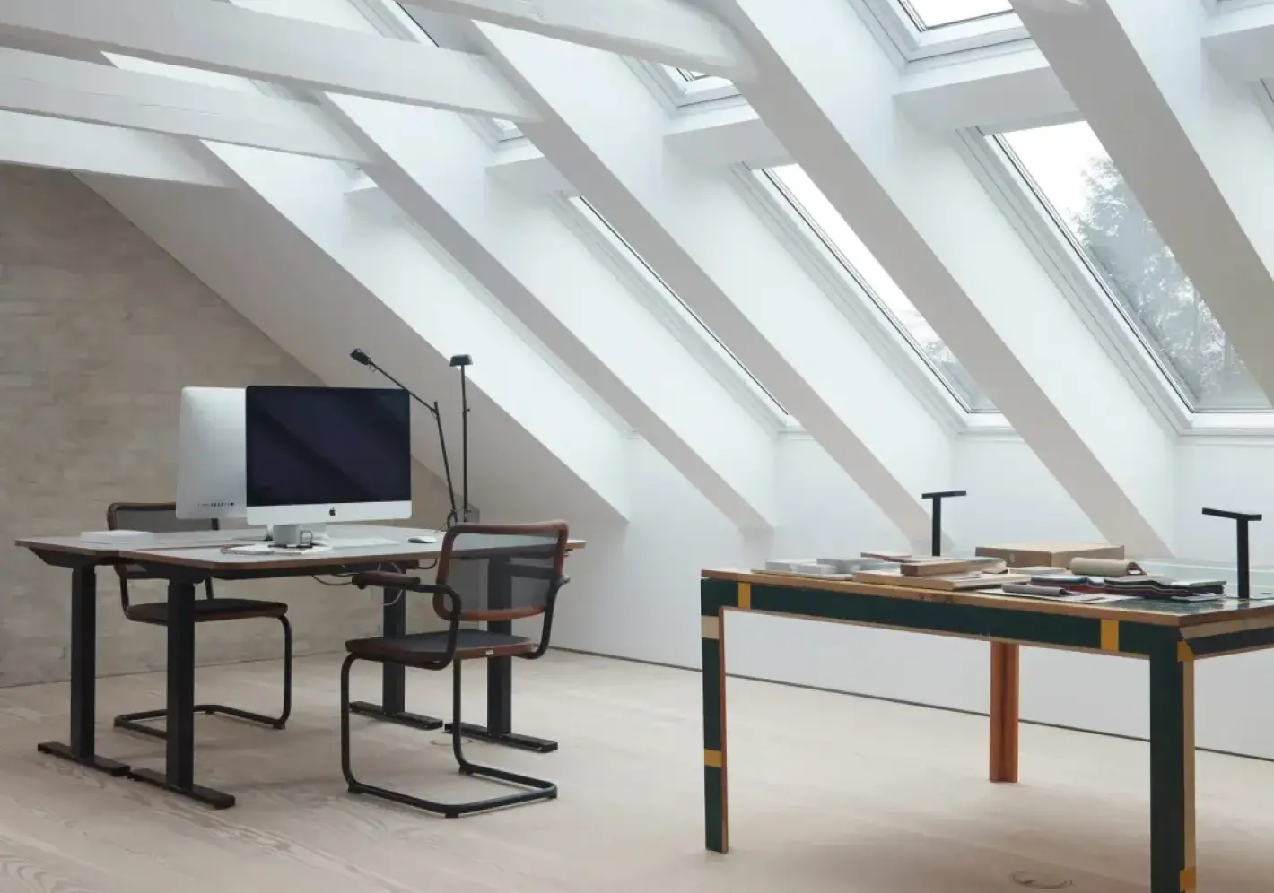 Modern attic office with slanted skylight windows, a desk with a computer, and another table with books and papers.