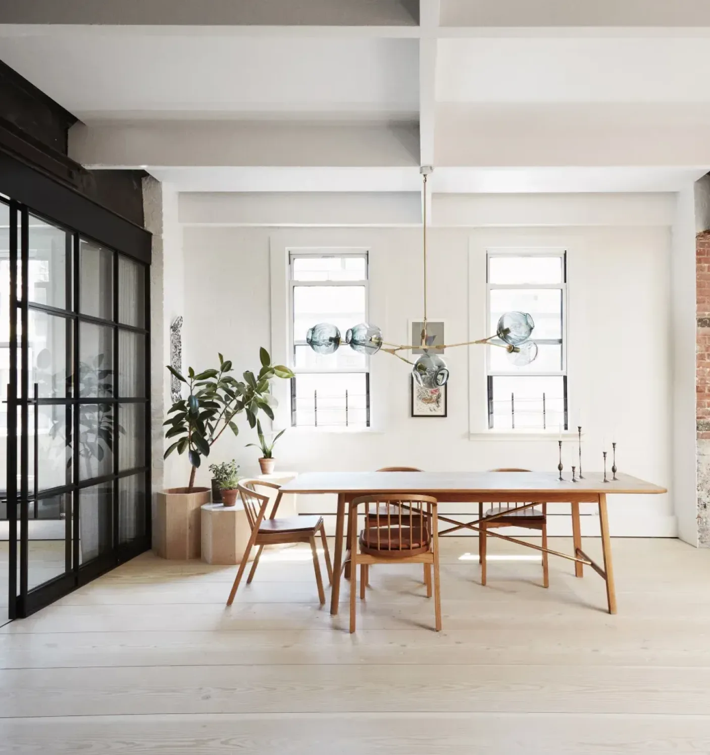 Modern dining room with a wooden table, four wooden chairs, hanging pendant light, and potted plants near two windows.