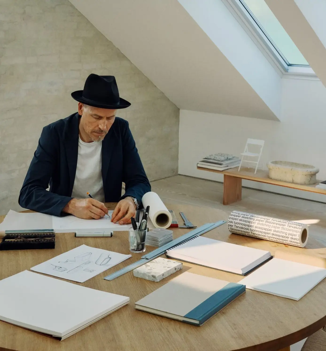 Man in a black hat and blazer sketching designs at a wooden table with drawing tools and sketchbooks in a bright room.