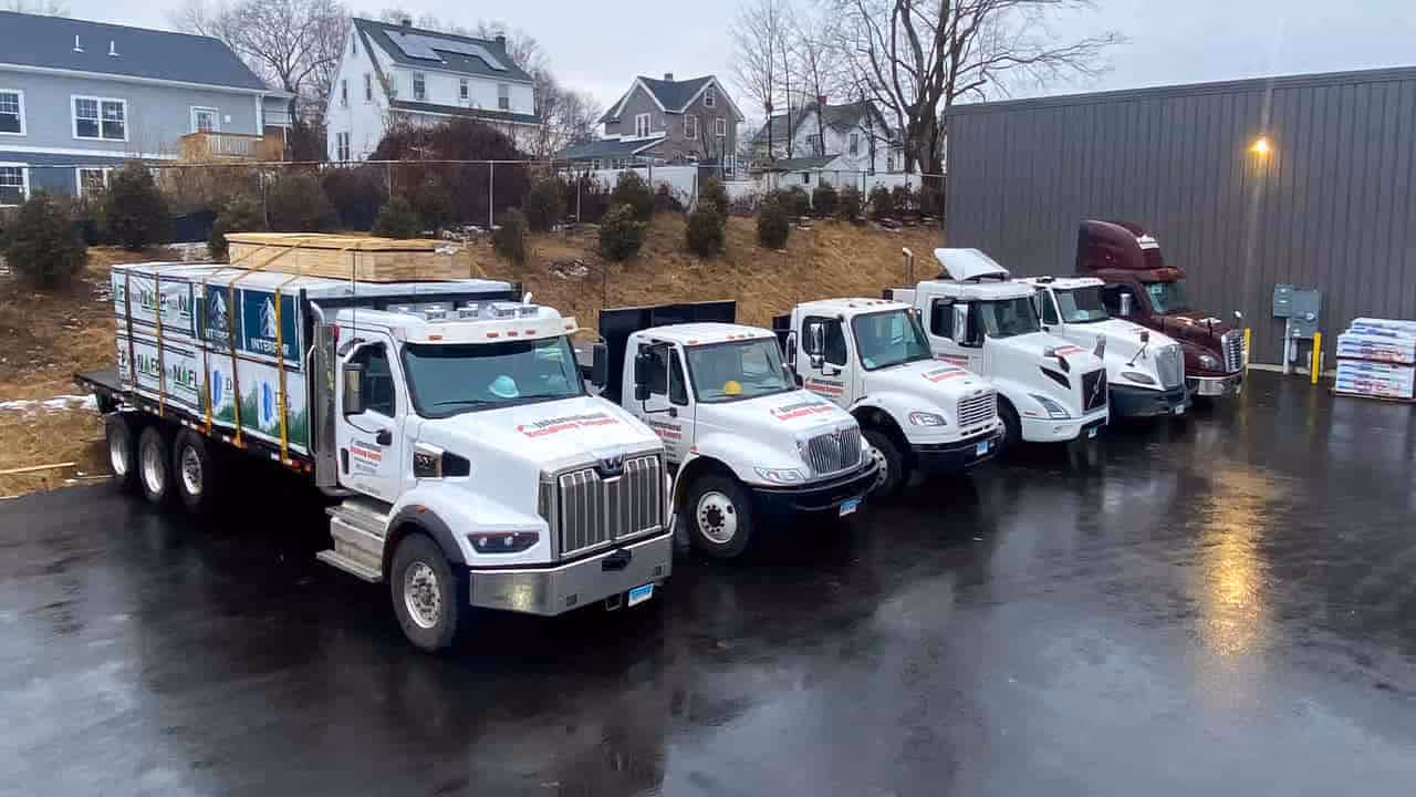 Fleet of five white delivery trucks, one loaded with lumber, parked on wet pavement outside industrial and residential buildings.