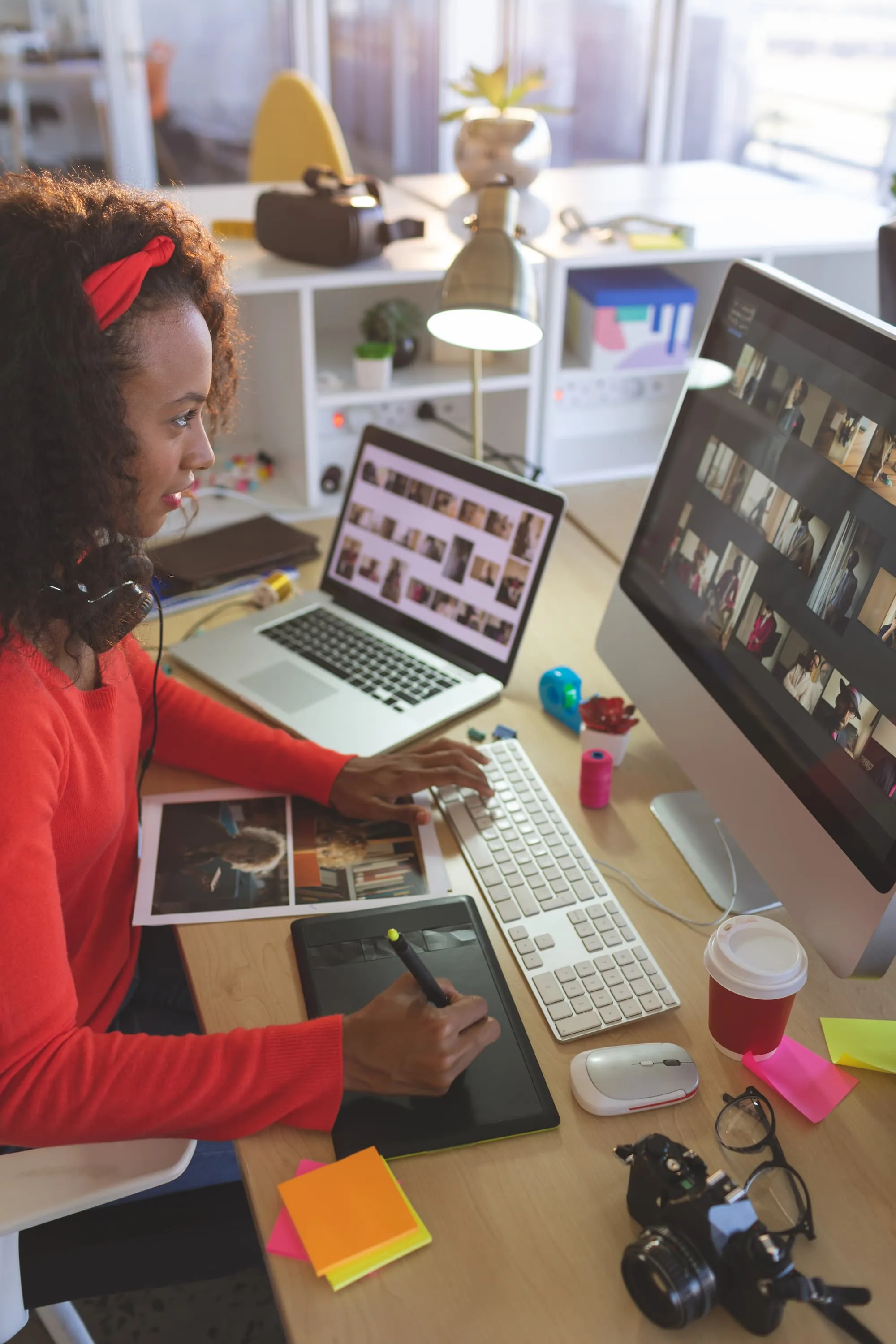 Woman editing photos at a desk with a laptop and desktop monitor displaying image thumbnails.