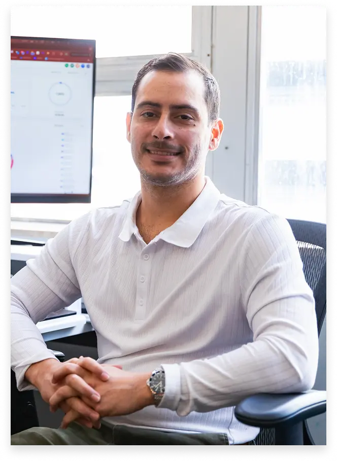 Man wearing a white long-sleeve polo shirt sitting in an office chair with hands clasped, smiling, with a computer monitor and window in the background.