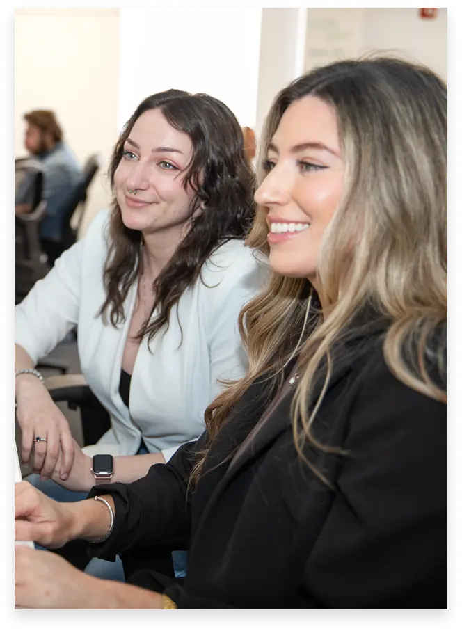 Two smiling women sitting closely, one with long wavy blonde hair wearing a black blazer, and the other with dark curly hair wearing a white jacket and a smartwatch, in a brightly lit indoor setting.