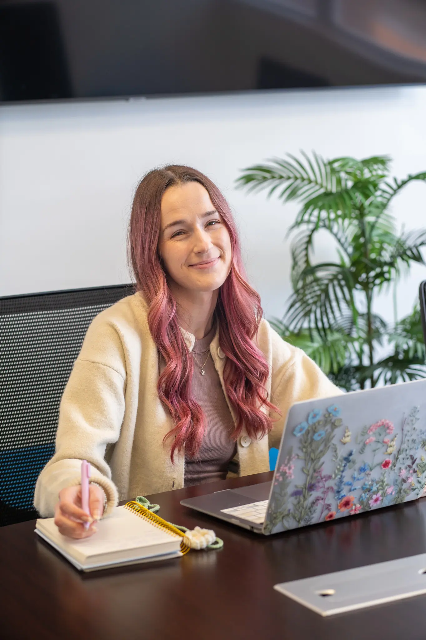 Smiling woman with long pink hair sitting at a desk, writing in a notebook with a pink pen, with a decorated laptop featuring floral designs in front of her and green plants in the background.