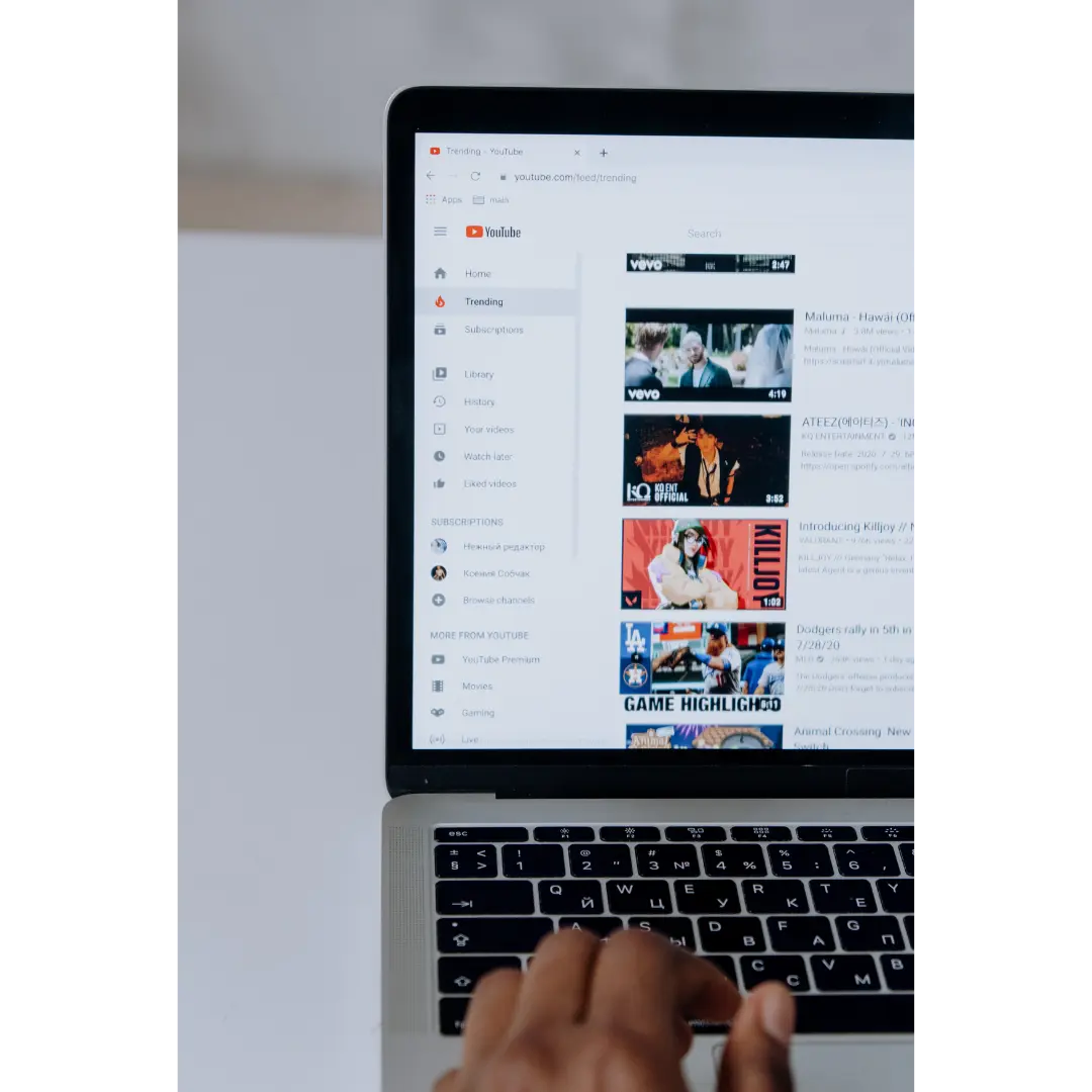 Person's hand on a laptop keyboard with the YouTube trending page open on the screen showing various video thumbnails and navigation menu.