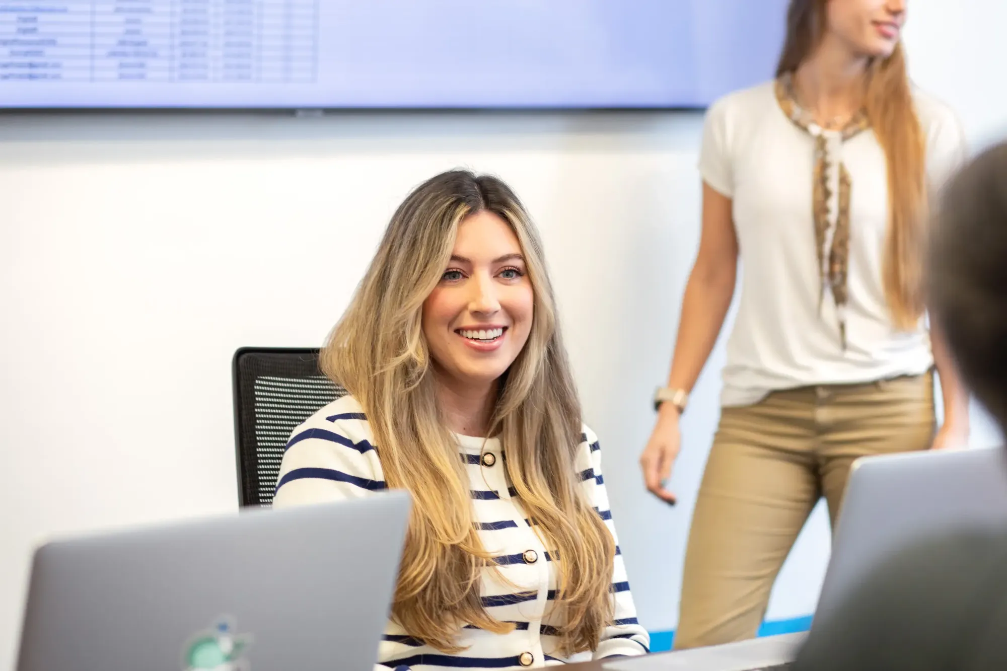 Smiling woman with long blonde hair in a striped top seated at a laptop during a team meeting, with a colleague standing beside a wall-mounted screen