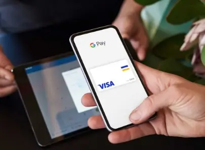 Mano sosteniendo un teléfono con Google Pay y una tarjeta Visa digital frente a una tableta.