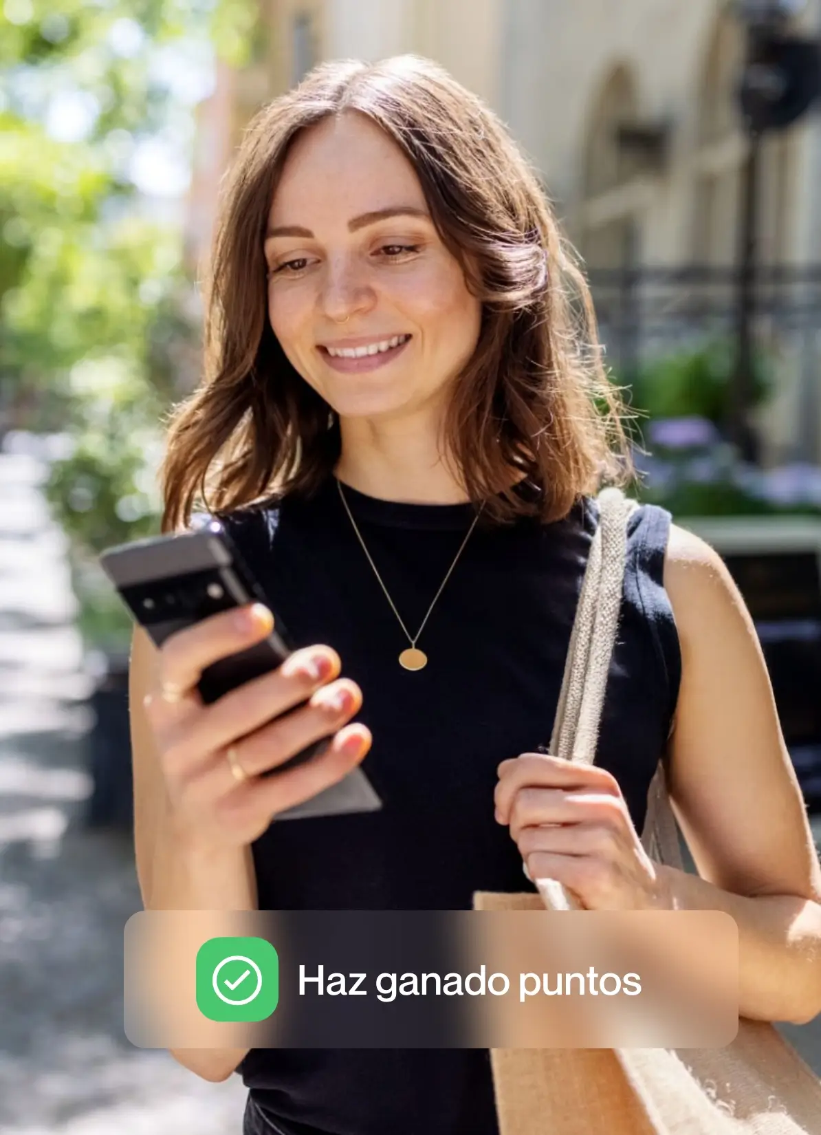 Mujer sonriente mirando su celular y cargando bolsa de tela en un día soleado.