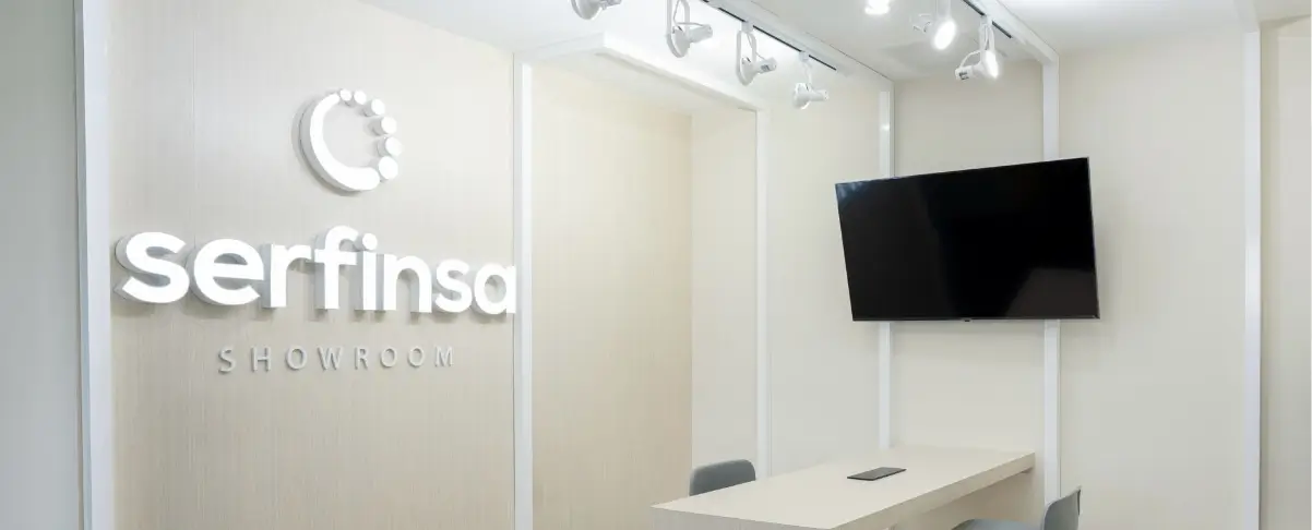 Interior de sala de exhibición con mesa, sillas, televisor y logotipo de Serfinsa en la pared.