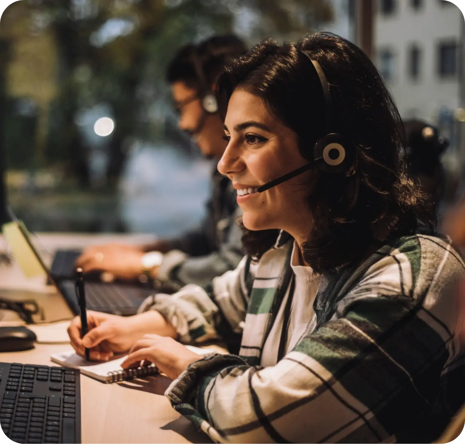 Mujer sonriente usando auriculares con micrófono y tomando notas frente a una computadora.