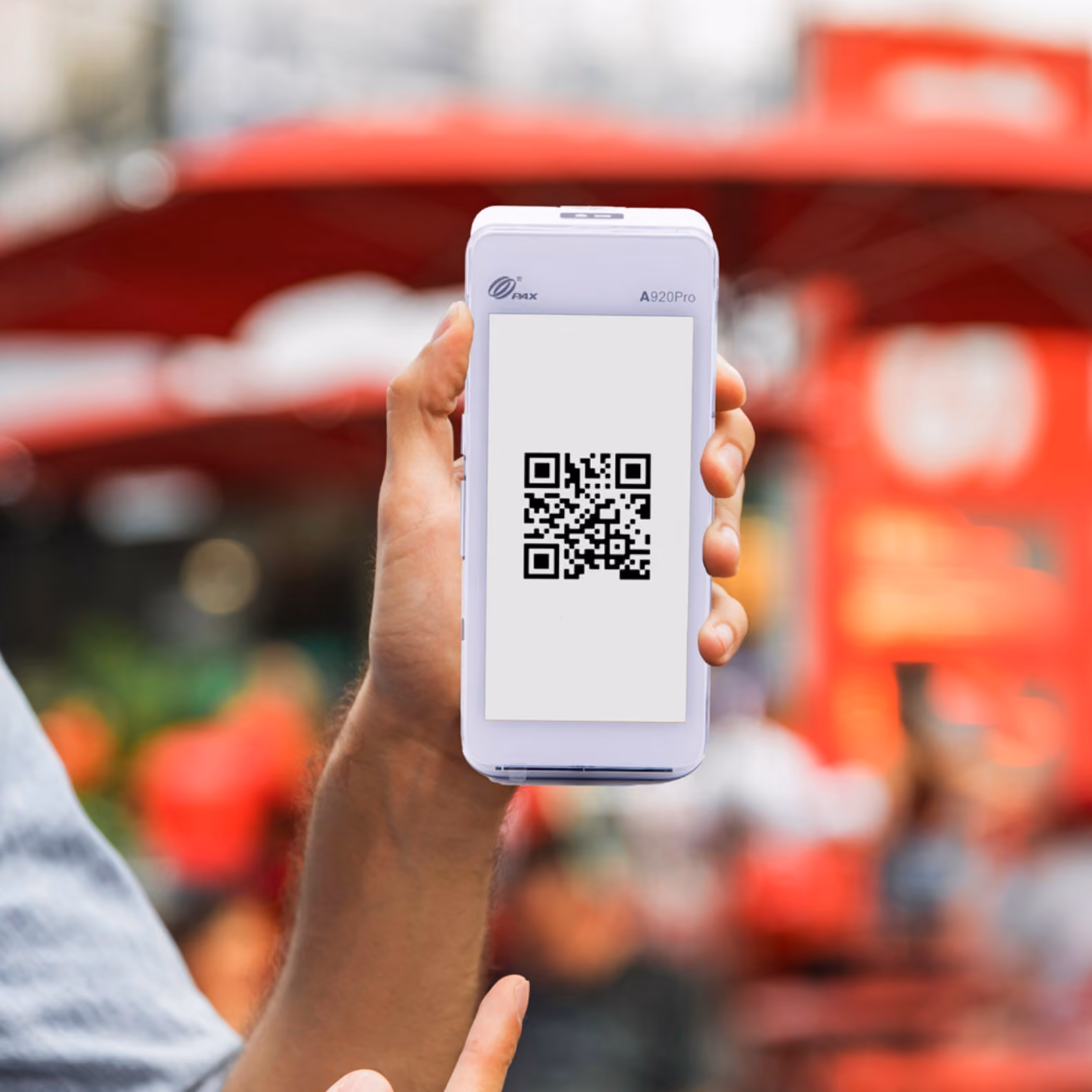 Mano sostiene un escáner de código QR blanco con un código en la pantalla en fondo urbano rojo desenfocado.