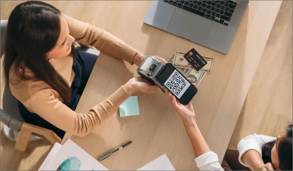 Dos mujeres usando pago móvil con código QR en dispositivo junto a dinero, tarjeta y laptop en mesa.