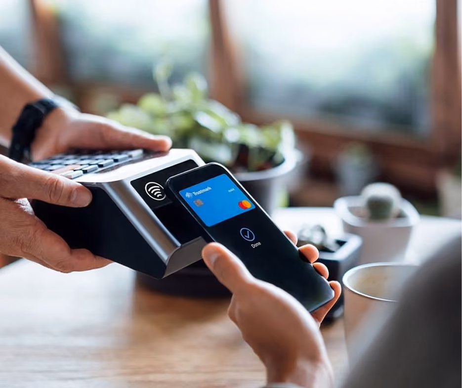 Persona pagando con un teléfono móvil usando una aplicación de tarjeta bancaria en un lector de tarjetas sin contacto en una mesa con plantas y una taza.