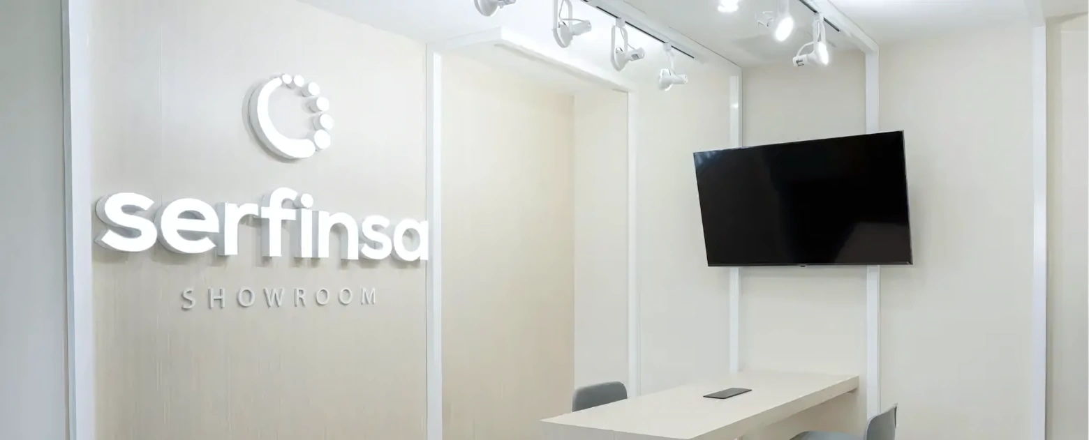 Interior de sala de exhibición con mesa, sillas, televisor y logotipo de Serfinsa en la pared.