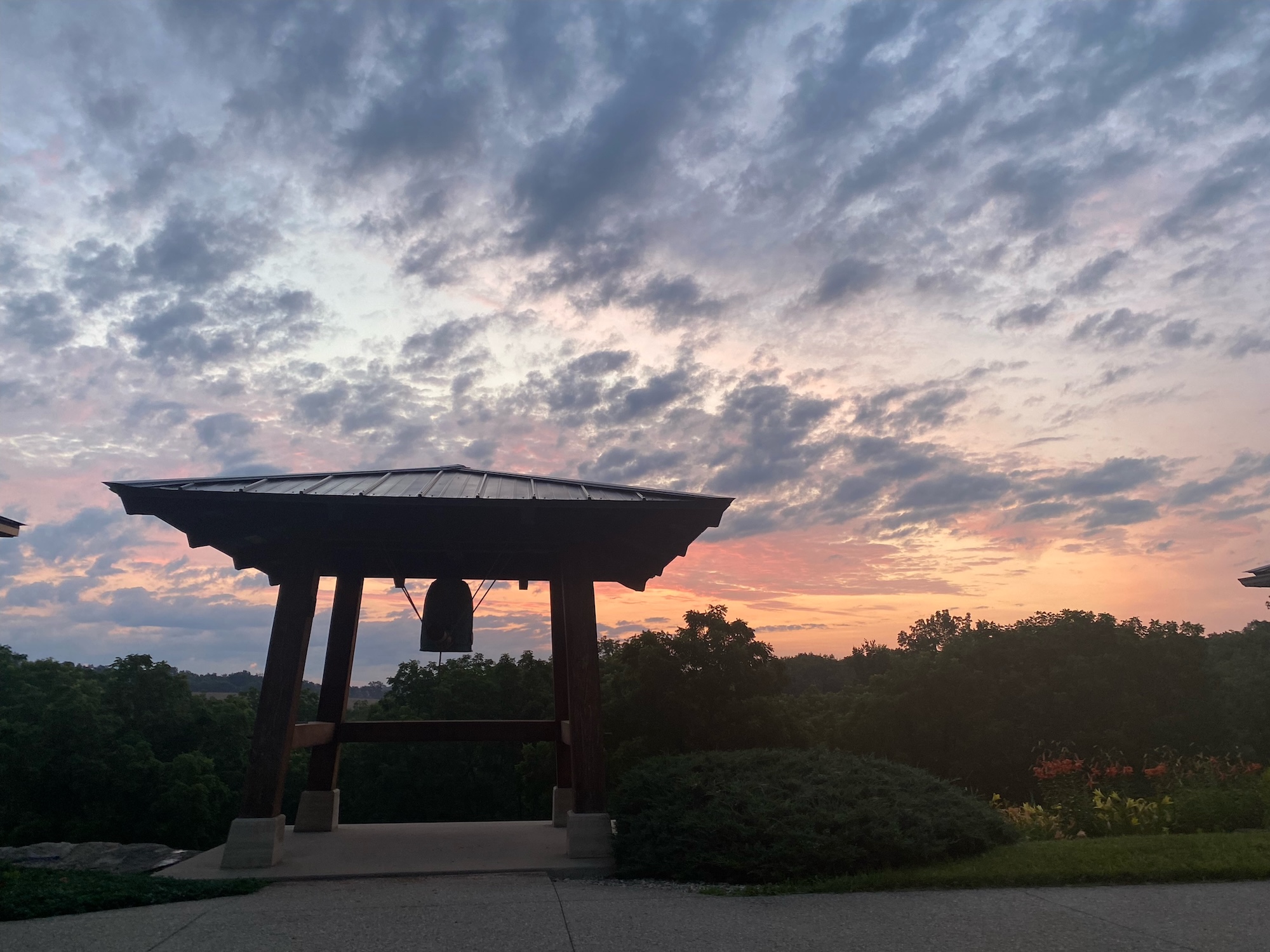 Wooden bell pavilion silhouetted against a colorful sunset sky with scattered clouds and surrounding greenery.
