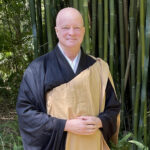 Smiling man dressed in traditional Buddhist monk robes standing in front of bamboo stalks.