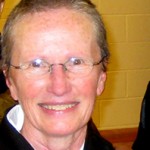 Smiling elderly woman with glasses and short light hair in front of a yellow wall.