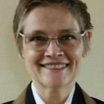 Smiling person with short hair, wearing glasses, a white shirt, and a brown jacket.