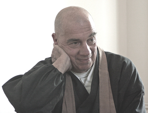 A man wearing traditional dark robes with a brown scarf, resting his head on his hand and smiling gently.