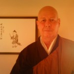 A man wearing traditional monk robes standing in front of a wall with Japanese calligraphy art.