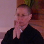 A person with short hair and glasses wearing a dark robe, holding hands together in a prayer or meditation gesture indoors.