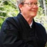 Smiling older person wearing glasses and a black jacket seated outdoors with green foliage background.