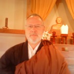 Portrait of a man wearing glasses and brown monk robes, sitting in a warmly lit room with religious statues and candles in the background.