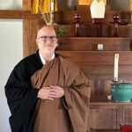 Smiling Buddhist monk wearing traditional robes standing in front of a wooden altar with candles and a green bowl.