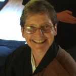 Smiling older person wearing glasses and a dark robe sitting indoors with a hand resting on their shoulder.
