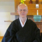 Portrait of an elderly man wearing traditional black robes sitting indoors with a wooden shelf and decorative items in the background.