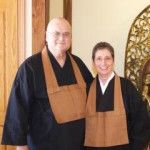 Two people wearing traditional monk robes standing indoors in front of a wooden door and decorative screen.