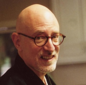 Smiling man with glasses and a short beard wearing a dark jacket indoors.