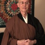 An older man wearing glasses and traditional brown and black meditation robes standing in front of a colorful tapestry.