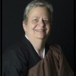 Smiling person with short gray hair wearing a dark robe and a brown drape over one shoulder against a black background.