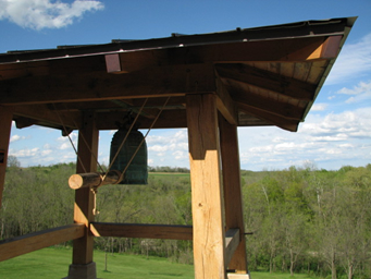 Wooden bell tower structure with a green bell hanging inside, set against a background of forest and blue sky with clouds.