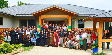 Large diverse group of people, including monks in traditional robes, gathered in front of a wooden building with a metal roof.