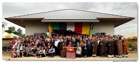 Large diverse group of people, including monks in robes, gathered in front of a building with colorful banners for a dedication event.
